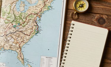 Clean, top-down professional photography of a wooden workspace featuring a topographical map of a North American trail, a vintage compass, and a field notebook. Earthy tones of sand and stone create a calm, focused atmosphere.