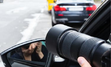 A private investigator uses a telephoto lens to photograph a car from a distance for surveillance.