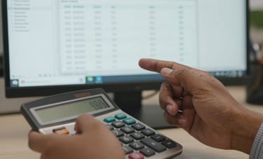 Close-up photography of a professional's hands using a calculator and reviewing financial spreadsheets on a computer screen in an Indian corporate office, focusing on precision and detail.