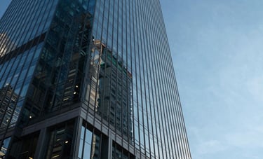 A low-angle shot of a modern glass skyscraper in a North American business district. The glass reflects a clear blue sky, with dark navy shadows and very light blue highlights, conveying a professional and high-trust atmosphere.
