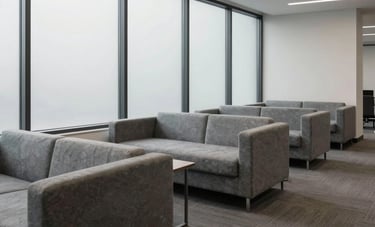 A minimalist North American / US corporate lounge area with modern slate gray furniture and large windows. The atmosphere is clean and approachable, emphasizing professional simplicity and a contemporary aesthetic.