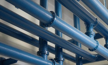 Professional architectural photography of commercial plumbing infrastructure in a North American / US office building. The pipes are neatly color-coded in shades of dark and light blue, showcasing organized industrial reliability and modern expertise.