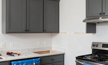 A mid-renovation photograph of a kitchen in a Los Angeles residence. New custom cabinets in dark gray are being aligned and installed against a fresh white wall. Steel blue protective film is visible on new stainless steel appliances. The atmosphere is productive and results-driven, showing the practical transformation process with professional tools nearby.