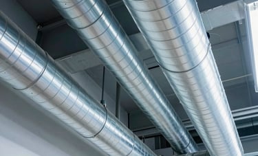 A wide-angle professional photograph of clean, shiny silver air ducts in a modern North American industrial ceiling. The lighting is bright white and blue, conveying a sense of sterile, clean airflow and technical excellence in ventilation services.