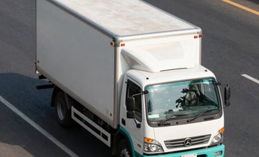A high-angle photograph of a modern commercial delivery truck with a white and teal exterior, driving on a clear highway near Delhi, India. The lighting is bright afternoon sun, suggesting speed and reliability.