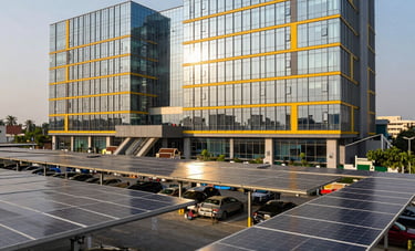 A wide professional shot of a modern glass-clad commercial office building in Lucknow, India, featuring an extensive solar canopy over the parking area. The lighting is bright afternoon sun, reflecting off solar yellow accents on the building's facade.