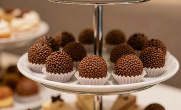 Close-up photography of a tiered dessert stand in a modern Brazilian cafe. It features delicate brigadeiros and slices of gourmet cake. Soft beige cream and light brown tones dominate the warm lighting.