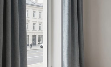 The interior of a bright, minimalist apartment in a Central European style. Through the window, a glimpse of a clean, modern street is visible. The scene is lit by soft daylight, emphasizing textures of greyish blue textiles and off-white walls, suggesting high-quality real estate services.