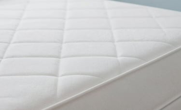 A professional close-up photograph of a bright, clean white mattress with crisp fabric texture. The setting is a minimalist bedroom with soft natural lighting and light blue accents. The image looks fresh, sterile, and inviting. South American home style.