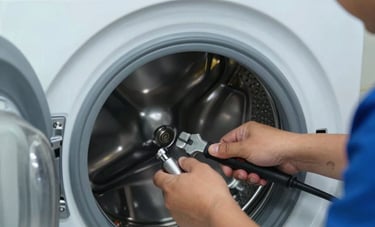 Close-up photography of a technician's skilled hands using professional tools to repair the drum of a modern washing machine. The setting is a clean, well-lit utility room in a Middle Eastern / Gulf home, featuring colors like light blue and grey-blue.