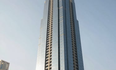 Professional architectural photography of a sleek high-rise residential tower in Ajman, Middle Eastern / Gulf metropolitan setting, bright morning light hitting the glass and mist-colored facade.
