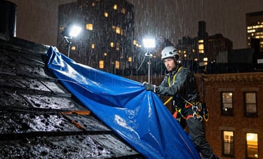 A dramatic North American night scene showing a professional roofer securing a heavy blue tarp on a storm-damaged roof in New York City. Rain is falling, and professional work lights illuminate the scene, conveying urgency.