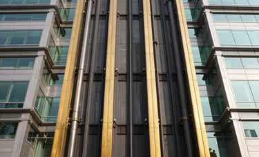 A wide architectural shot of a grand South Asian government headquarters featuring high-tech glass-walled elevators. The composition is symmetrical and powerful, showcasing industrial sophistication. Sunlight glints off gold-colored steel frames and polished black surfaces.