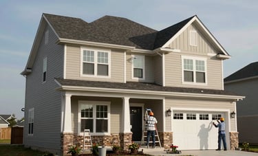An exterior shot of a large, two-story North American suburban home. Professional painters are using ladders to carefully apply beige and black accents to the siding and trim under a clear sky.