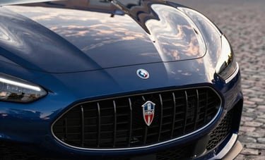 Close-up photography of a dark blue Italian sports car's front grille and emblem, reflecting a sunset sky. The car is situated in a cobblestone driveway of a luxury estate in the North American / European Luxury Automotive Market, evoking exclusivity and precision.