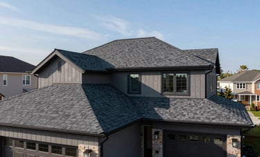 A wide angle shot of a beautifully restored modern home in a North American neighborhood, featuring a brand-new roof with deep charcoal gray shingles under a clear sky, emphasizing high-end craftsmanship and curb appeal.
