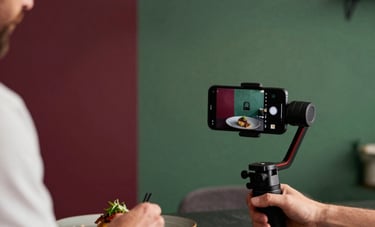Behind-the-scenes shot of a content creator in a Northern European setting, using a gimbal and smartphone to film a chef plating a dish. Modern, artisanal aesthetic with Matte Forest Green and Deep Ripe Crimson accents in the decor.