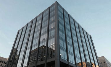 The exterior of a sleek, modern glass office building in a North American business district. The shot is a low-angle perspective against a clear sky, showcasing the sophisticated dark gray and muted blue reflections of the architecture.