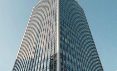 An architectural photograph of a sleek, contemporary glass office tower in a North American business district. The shot is taken from a low angle against a clear blue sky, emphasizing corporate success. Reflective surfaces show light blue and off-white tones.