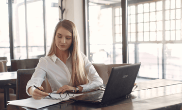 a woman sitting at a table with a laptop, notebook, pen and glasses