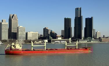 A red oceangoing ship with tan deck cranes passing downtown Detroit.