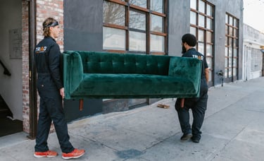 Professional movers carrying a green velvet sofa out of a building on a city sidewalk.