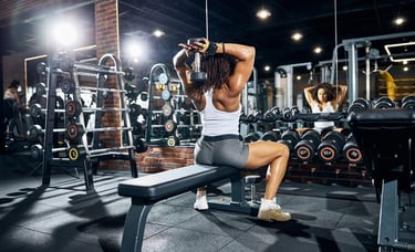 A muscular woman performs overhead dumbbell triceps extensions on a gym bench.