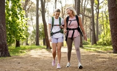 Two happy women with backpacks hiking on a sunny trail through a lush pine forest.