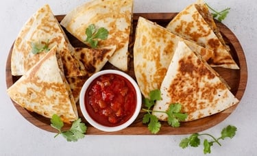 A wooden platter filled with toasted cheese and meat quesadilla triangles, served with fresh salsa and cilantro.