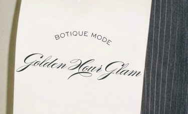 Mano sosteniendo una invitación Golden Hour Glam de Botique Mode sobre un pantalón de raya.