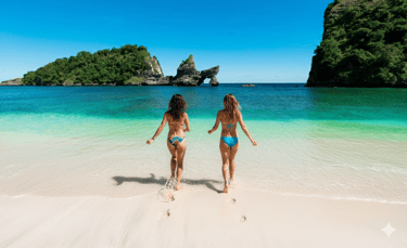 two women walking into turquoise ocean on a white sandy beach with limestone cliffs and lush green islands under blue sky