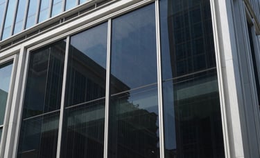 Photography of a modern glass building in an Indonesian city during a bright day, showing professional heat-rejecting window tinting being applied to large glass panels. The scene is clean, reflecting a blue sky, with a focus on the sleek dark tint finish.