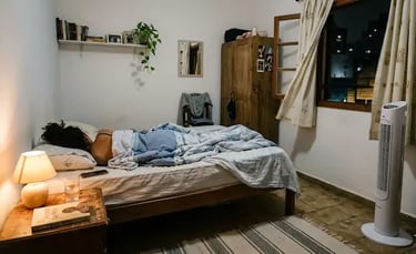 A person sleeping in a cozy bedroom with a warm bedside lamp and a tall white tower fan.