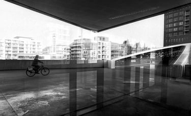 Black and white street photography of a cyclist riding past modern urban architecture and reflections.