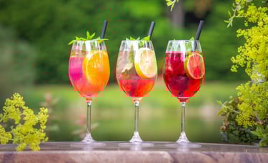 Drei lebendige Sommer-Cocktails in hohen Stielgläsern auf rustikalem Holz, garniert mit Früchte