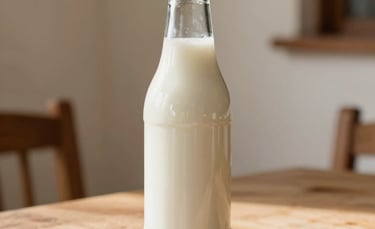 A minimalist, close-up photograph of a clean glass bottle filled with creamy white kefir, placed on a rustic light wood table. The background is a soft cream wall in a bright Latin American home. Natural morning sunlight creates gentle shadows, emphasizing the smooth texture of the liquid.
