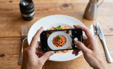 A top-down professional photography shot of a rustic wooden restaurant table in a Western European setting. A person is capturing a photo of a vibrant pasta dish using a smartphone, surrounded by natural morning light and cozy Scandinavian decor.