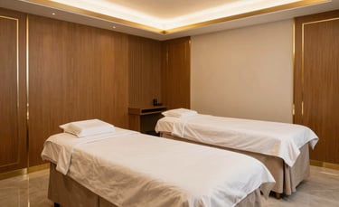 A wide-angle professional photograph of a luxury spa suite in a South Asian boutique hotel. Modern Indian interior design with gold-trimmed wooden panels and a soft light beige aesthetic. Large comfortable massage beds are neatly made with white linens.