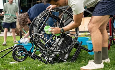A wheelchair flipped upside down on the grass with someone sponging the wheels