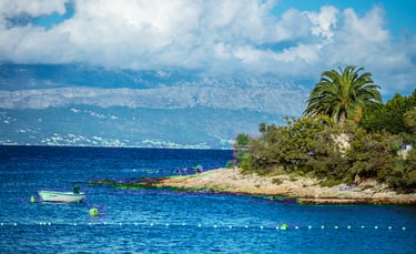 Small boat anchored in a blue Mediterranean bay with rocky shores and palm trees against a mountain backdrop.