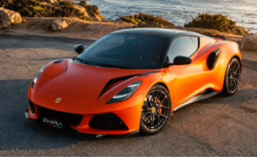 Lotus Emira 420 Sport laranja parado em estrada costeira ao pôr do sol.