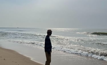 Man in a straw hat walking along a sunny beach with ocean waves crashing on the shore.