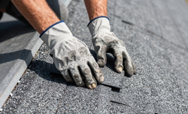 A close-up photograph of a professional roofer's hands in the United States, wearing protective gloves, repairing an asphalt shingle roof. The lighting is bright and clear daylight, emphasizing the clean texture of the grey materials and the precision of the work.