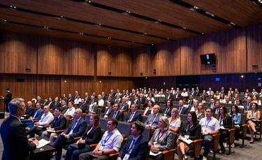 A wide-angle professional photograph of a large, modern auditorium in a Brazilian convention center. The room is filled with an attentive audience listening to a speaker. The lighting is sophisticated, combining warm brown and dark blue tones, reflecting a highly organized and professional corporate atmosphere.