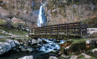 A wooden footbridge crossing a rocky stream with the Aber Falls waterfall in the background.