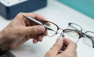A close-up photograph of hands working with precision tools on a pair of glasses in a professional optical setting, bright teal and navy accents in the room.