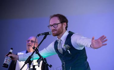 Groom in a green plaid waistcoat gives an energetic wedding speech holding a beer bottle at a microphone.