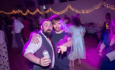 Two friends smiling and dancing under purple event lights at a wedding reception party with string lights.