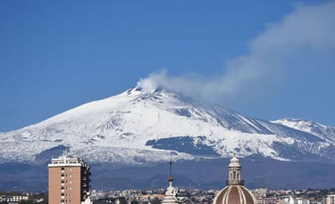 Panoramic view of snow-capped Mount Etna volcano emitting smoke above the skyline of Catania, Sicily.