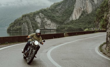 A person riding a vintage motorcycle on a winding coastal road with scenic mountain views.